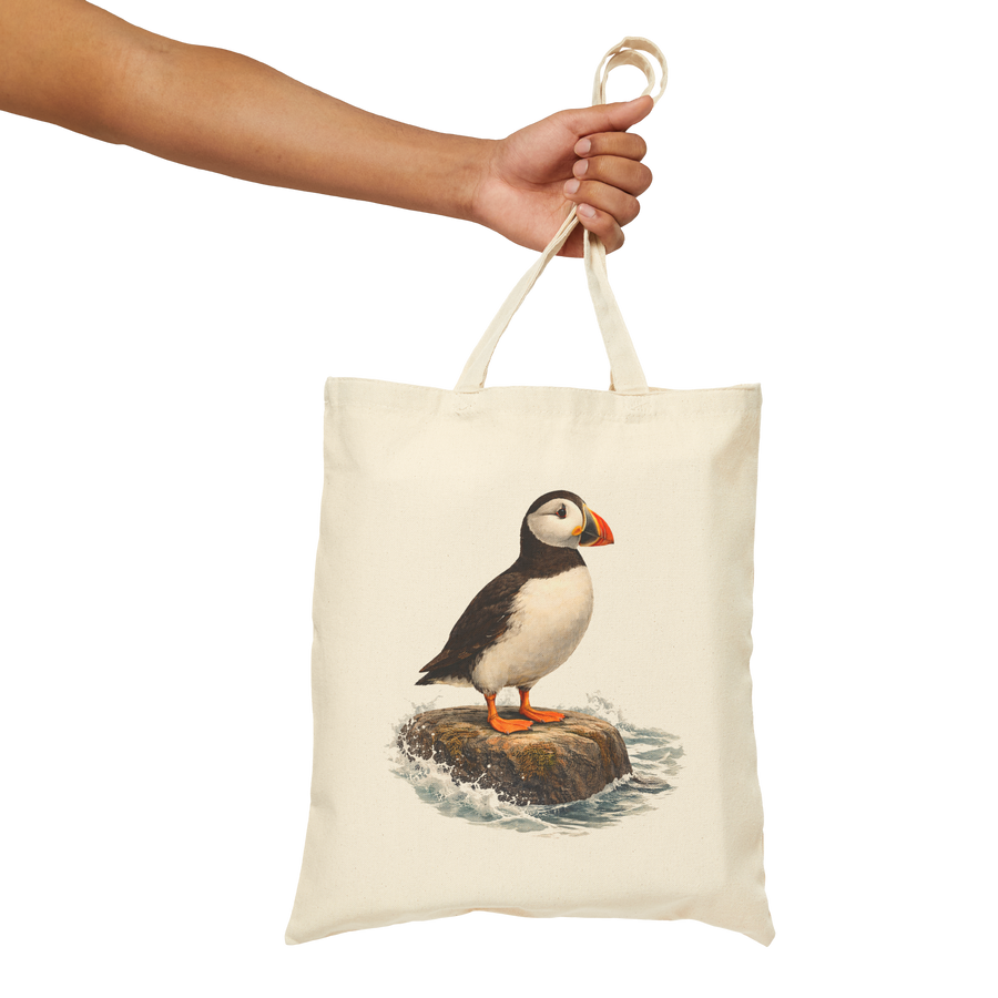 Natural cotton canvas tote bag held by its handles, printed with a puffin standing on a coastal rock